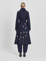 Load image into Gallery viewer, A model viewed from the back wearing a navy blue wool-blend coat with white embroidered poppy flowers, paired with matching navy trousers and black boots, against a light gray studio background.
