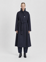 Load image into Gallery viewer, A model stands against a light gray studio background wearing a long dark navy wrap coat with a high collar, matching dark trousers, and black ankle boots.
